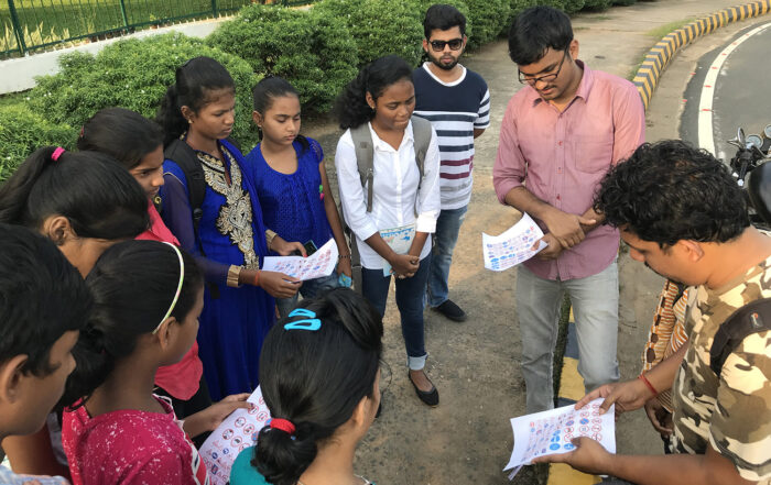 Two men presenting survey forms to a group of children on the footpath of a road for a safety-audit.