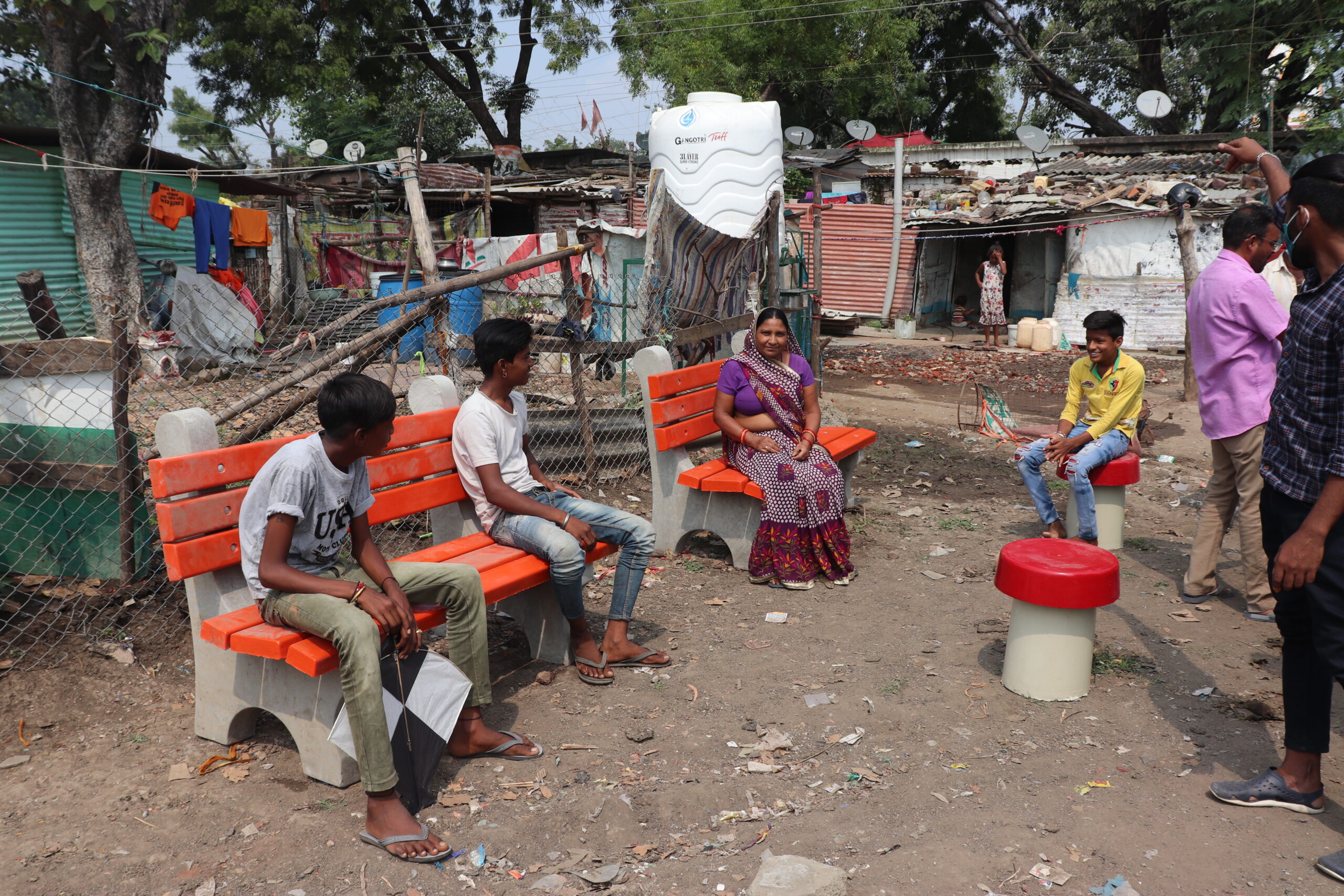Few boys and a lady are seated on an arrangement of benches and stools in the public area of a rural settlement. They are following the norms of social-distancing set during the COVID-19 pandemic.